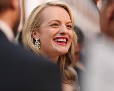 Elisabeth Moss smiling at a public event, wearing pearl earrings and red lipstick.