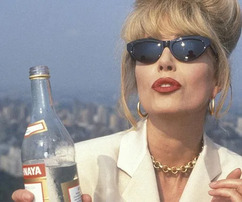 Woman with blond hair in sunglasses, red lipstick, white suit holding a bottle.