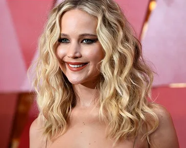 Jennifer Lawrence at a red carpet event, with long wavy blonde hair, wearing a strapless dress and smiling.