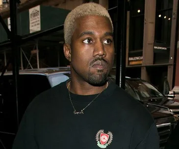 Man wearing dark shirt with emblem and gold necklace, standing outside in city with blonde hair.