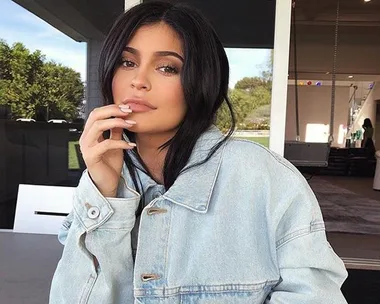 Woman with long dark hair wearing a light denim jacket sits outdoors, resting her chin lightly on her hand.