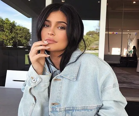 Woman with long dark hair wearing a light denim jacket sits outdoors, resting her chin lightly on her hand.