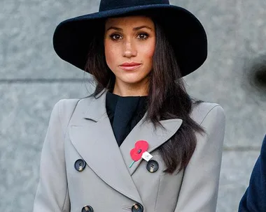 Woman in a grey coat and black wide-brimmed hat with a red poppy pin.