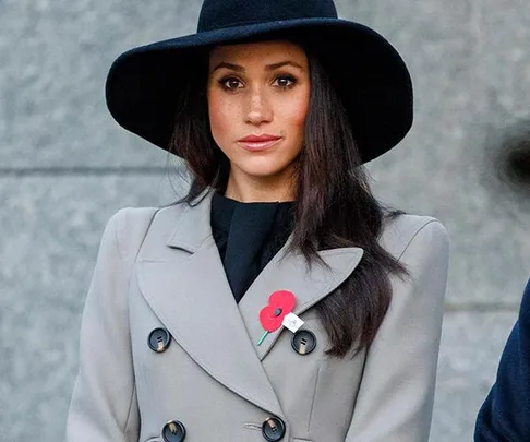 Woman in a grey coat and black wide-brimmed hat with a red poppy pin.