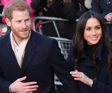 Prince Harry and Meghan Markle smiling and holding hands while walking outdoors.