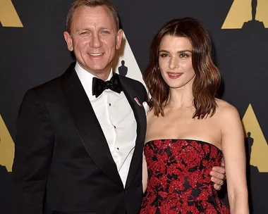 A man in a black tuxedo and a woman in a red and black strapless gown posing together at an event with an Oscars backdrop.