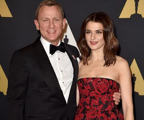 A man in a black tuxedo and a woman in a red and black strapless gown posing together at an event with an Oscars backdrop.