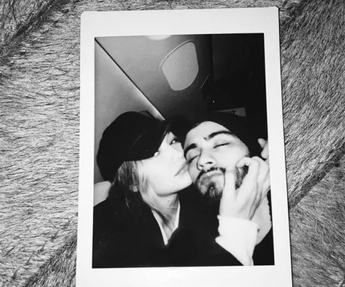 A black-and-white Polaroid of a couple sitting closely, the woman wearing a hat and kissing the man's cheek affectionately.