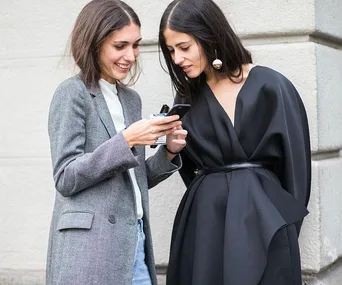 Two women smiling and looking at a smartphone, one in a grey blazer and the other in a black outfit with statement earrings.