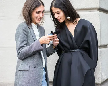 Two women smiling and looking at a smartphone, one in a grey blazer and the other in a black outfit with statement earrings.