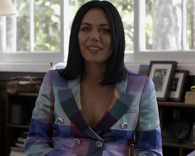 Woman in colorful checkered blazer, sitting and smiling in well-lit room with framed photo and greenery outside window.