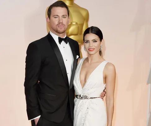 A couple dressed in elegant formal wear posing together, with an Oscar statue in the background.