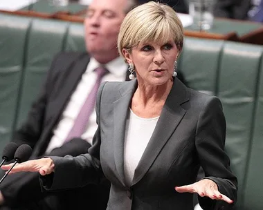 A female politician speaks emphatically at a government assembly while a male colleague appears inattentive in the background.