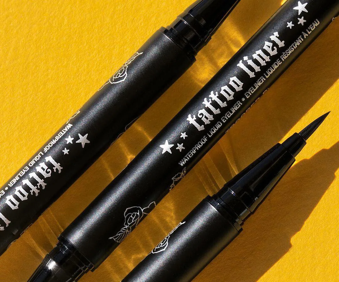 Close-up of three black Kat Von D Tattoo Liner pens on a yellow background with detailed tips and branding.