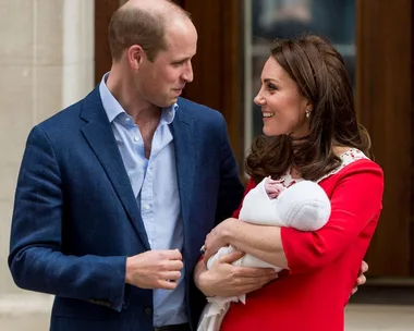 A couple smiling at each other while the woman holds a newborn baby wrapped in a white blanket.