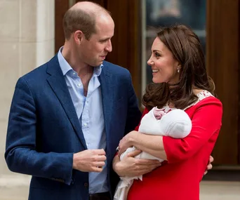 Royal couple smiling and looking at each other while holding their newborn baby outside a building.