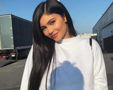 A woman with long, dark hair and a white sweatshirt smiles outdoors with trucks and buildings in the background.