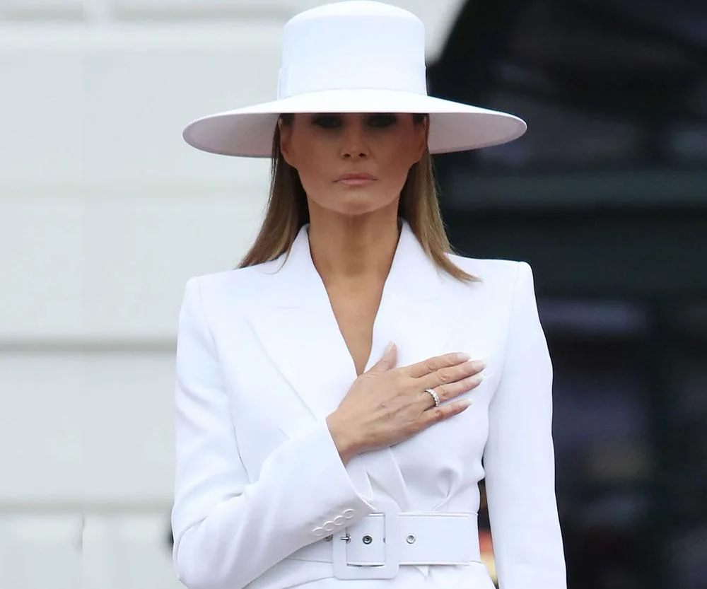 A woman dressed in a white suit and wide-brimmed hat with her hand over her heart.