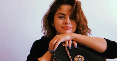 A woman with wavy hair and blue nail polish holding a black handbag, smiling with a white background.