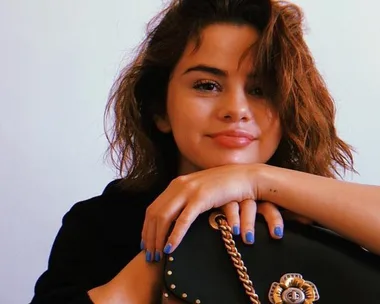 A woman with wavy hair and blue nail polish holding a black handbag, smiling with a white background.