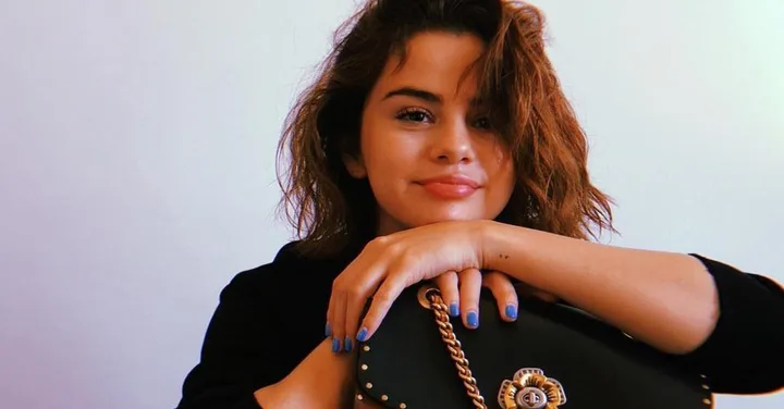 A woman with wavy hair and blue nail polish holding a black handbag, smiling with a white background.