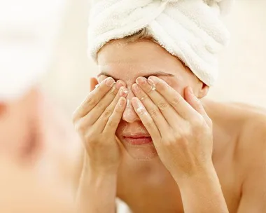 A woman with a towel on her head covers her eyes with her hands while washing her face.