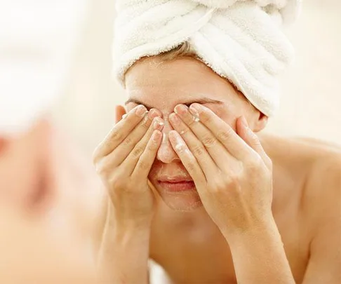 A woman with a towel on her head covers her eyes with her hands while washing her face.