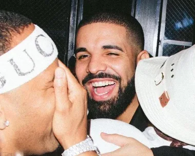 Drake smiling at a party, surrounded by friends wearing a Gucci headband and a white hat.