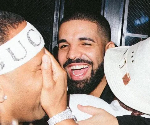 Drake smiling at a party, surrounded by friends wearing a Gucci headband and a white hat.