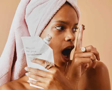 Woman with towel on head holding a packet, looking at chocolate substance on finger, surprised expression.