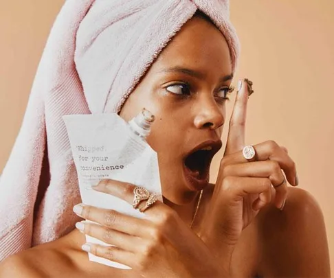 Woman with towel on head holding a packet, looking at chocolate substance on finger, surprised expression.