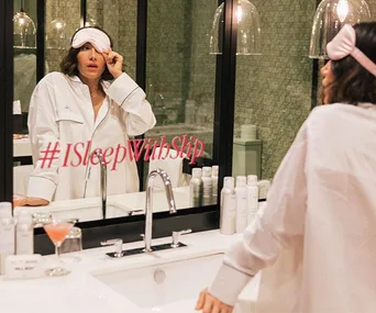 Woman in pajamas lifts pink eye mask in bathroom, hashtag #ISleepWithSlip on mirror, toiletries and cocktail on counter.