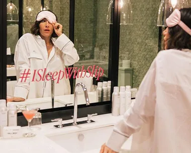 Woman in pajamas lifts pink eye mask in bathroom, hashtag #ISleepWithSlip on mirror, toiletries and cocktail on counter.