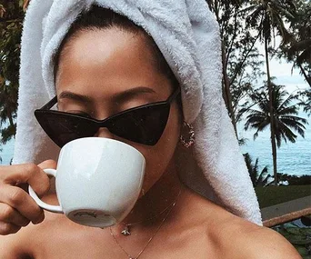 Person with a towel on head and sunglasses drinking from a white mug with tropical trees in the background.