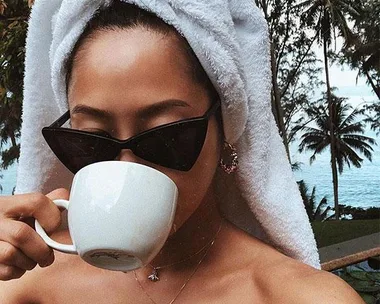 Person with a towel on head and sunglasses drinking from a white mug with tropical trees in the background.