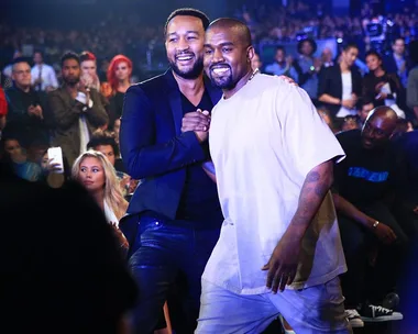 Two men are smiling and shaking hands at an event, surrounded by a crowd.