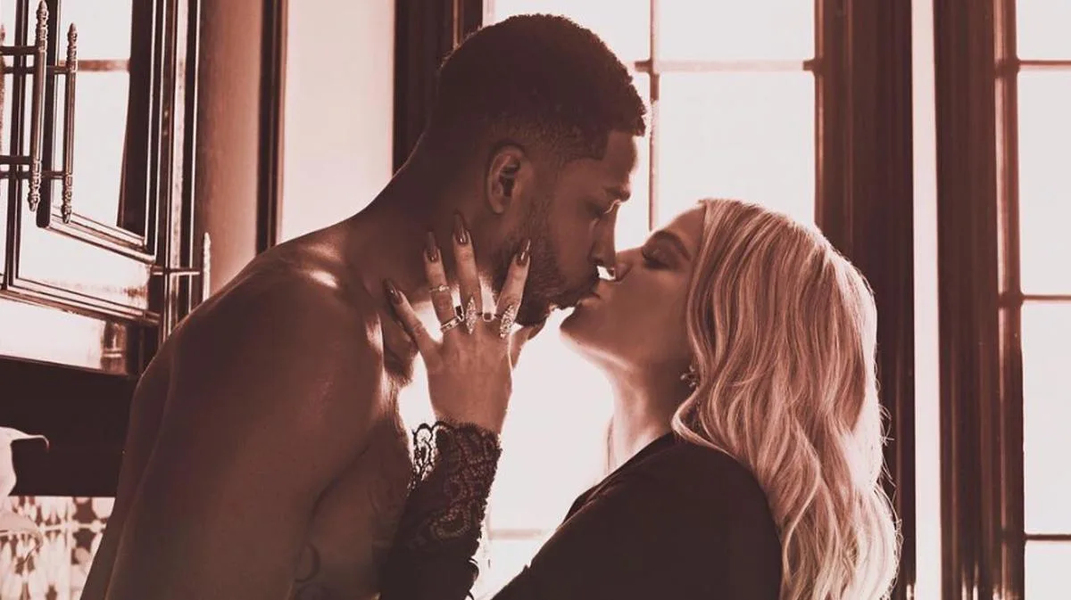 A couple sharing a kiss in a warmly lit room, with one person shirtless and the other dressed in black, with soft window light in the background.