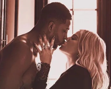 A couple sharing a kiss in a warmly lit room, with one person shirtless and the other dressed in black, with soft window light in the background.