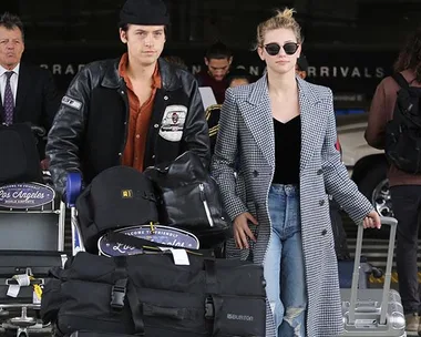 Two individuals with luggage carts at an airport terminal, wearing casual travel outfits and sunglasses.