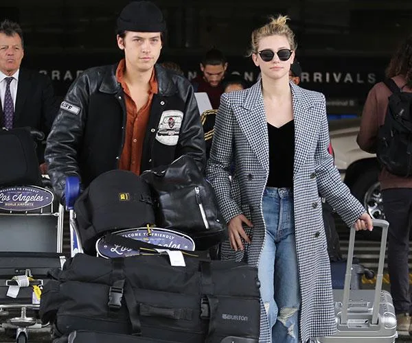 Two individuals with luggage carts at an airport terminal, wearing casual travel outfits and sunglasses.