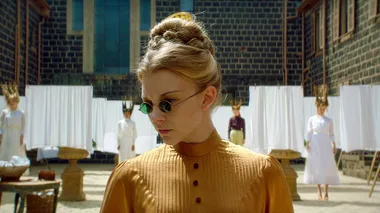 A woman in vintage costume with sunglasses stands in a courtyard, with three women in white dresses in the background.