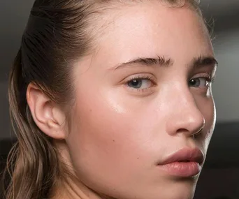 Young person with clear, glowing skin and slicked back hair, showcasing dewy complexion in a beauty setting.