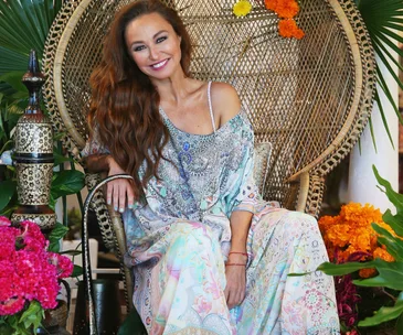 A woman in colorful attire sits on a wicker chair surrounded by vibrant flowers and foliage, smiling at the camera.