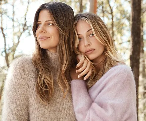 Two women in soft, cozy sweaters embrace in a serene forest setting, one smiling and the other resting her head on her hand.