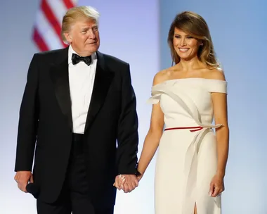 A man in a tuxedo holds hands with a woman in an elegant white dress, with an American flag in the background.