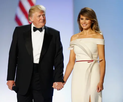 A man in a tuxedo holds hands with a woman in an elegant white dress, with an American flag in the background.