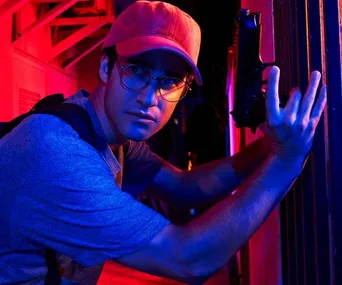 Darren Criss as Andrew Cunanan, wearing glasses and a red cap, holding a gun, in "The Assassination of Gianni Versace".