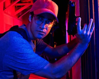 Darren Criss as Andrew Cunanan, wearing glasses and a red cap, holding a gun, in "The Assassination of Gianni Versace".