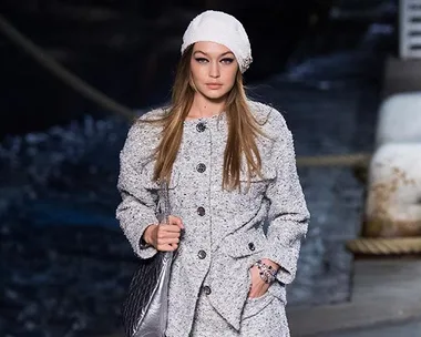 Model Gigi Hadid wearing a white outfit and hat, walking on a runway for a fashion show.