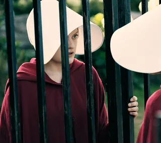 A woman in a white bonnet and red cloak stands behind black bars in a scene from The Handmaid's Tale.